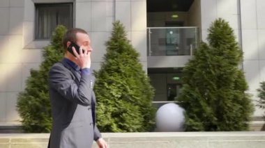 Genç bir işadamı sokakta yürürken mutlu bir telefon görüşmesi iletişim kurar. Takım elbiseli neşeli bir adam akıllı telefonda konuşurken gülümsüyor. Arka planda küçük ağaçlar var. 4k görüntü.