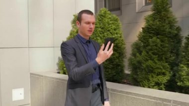 Genç bir işadamı sokakta yürüyor ve görüntülü görüşme hakkında konuşuyor. Klasik takım elbiseli genç bir adam video konferansında bir konuşma açar. Arka planda küçük yeşil ağaçlar. 4k görüntü.