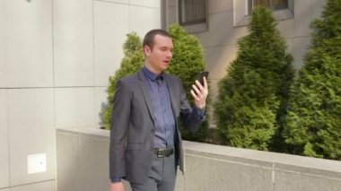 Genç bir işadamı sokakta yürüyor ve görüntülü görüşme hakkında konuşuyor. Klasik takım elbiseli genç bir adam video konferansında bir konuşma açar. Arka planda küçük yeşil ağaçlar. 4k görüntü.