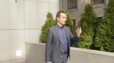 Genç bir işadamı sokakta yürüyor ve görüntülü görüşme hakkında konuşuyor. Klasik takım elbiseli genç bir adam video konferansında bir konuşma açar. Arka planda küçük yeşil ağaçlar. 4k görüntü.