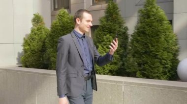Genç bir işadamı sokakta yürürken ve mutlu görüntülü arama iletişim kurar. Klasik takım elbiseli neşeli bir adam video konferansında konuşurken gülümsüyor. 4k görüntü.