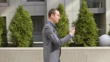 Genç bir işadamı sokakta yürüyor ve görüntülü görüşme hakkında konuşuyor. Klasik takım elbiseli genç bir adam video konferansında bir konuşma açar. Arka planda küçük yeşil ağaçlar. 4k görüntü.