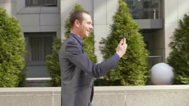 Genç bir işadamı sokakta yürürken ve mutlu görüntülü arama iletişim kurar. Klasik takım elbiseli neşeli bir adam video konferansında konuşurken gülümsüyor. 4k görüntü.