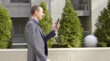 Genç bir işadamı sokakta yürürken ve mutlu görüntülü arama iletişim kurar. Klasik takım elbiseli neşeli bir adam video konferansında konuşurken gülümsüyor. 4k görüntü.
