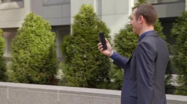 Genç bir işadamı sokakta yürürken ve mutlu görüntülü arama iletişim kurar. Klasik takım elbiseli neşeli bir adam video konferansında konuşurken gülümsüyor. 4k görüntü.