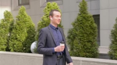 Kablosuz kulaklıkla yürüyen ve mutlu bir telefon görüşmesi yle iletişim kuranan genç bir işadamı. Elinde bir bardak kahve ile neşeli bir adam konuşurken gülümsüyor. 4k görüntü.