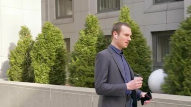 Genç işadamı kablosuz kulaklıkla yürüyor ve telefonda agresif bir tartışma yürütüyor. Elinde bir bardak kahve olan bir adam telefonda konuşarak öfkesini gösterir. 4k görüntü.