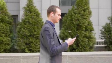 Genç işadamı kulaklıkları kablosuz kulaklıkla sokakta yürüyor ve telefona bir mesaj yazıyor. Klasik takım elbiseli adam kahve içer ve akıllı telefon kullanır. 4k görüntü