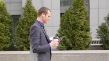 Genç işadamı kulaklıkları kablosuz kulaklıkla sokakta yürüyor ve telefona bir mesaj yazıyor. Klasik takım elbiseli adam kahve içer ve akıllı telefon kullanır. 4k görüntü