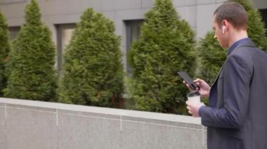Genç işadamı kulaklıkları kablosuz kulaklıkla sokakta yürüyor ve telefona bir mesaj yazıyor. Klasik takım elbiseli adam kahve içer ve akıllı telefon kullanır. 4k görüntü.