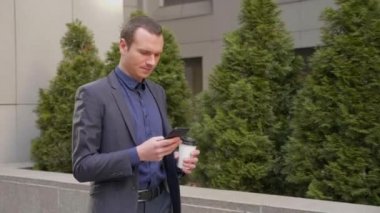 Elinde bir bardak kahveyle caddede yürüyen genç işadamı telefona bir mesaj yazıyor. Klasik takım elbiseli adam kahve içer ve akıllı telefon kullanır. 4k görüntü.