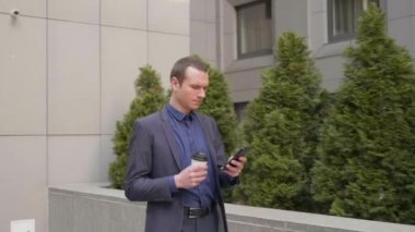 Elinde bir bardak kahveyle caddede yürüyen genç işadamı telefona bir mesaj yazıyor. Klasik takım elbiseli adam kahve içer ve akıllı telefon kullanır. 4k görüntü