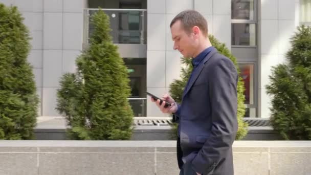 若いビジネスマンが通りを歩いてスマートフォンにメッセージを書きます 古典的なスーツの男は自分の携帯電話を使う 背景に小さな緑の木 4K映像 — ストック動画