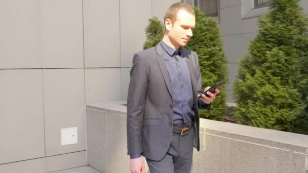 若いビジネスマンが通りを歩いてスマートフォンにメッセージを書きます 古典的なスーツの男は自分の電話を使う 背景に小さな緑の木 4K映像 — ストック動画