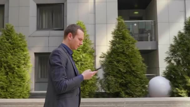 年轻的商人走在街上 用智能手机留言 穿着经典西服的男人用自己的电话 背景上的绿色小树 4K镜头 — 图库视频影像