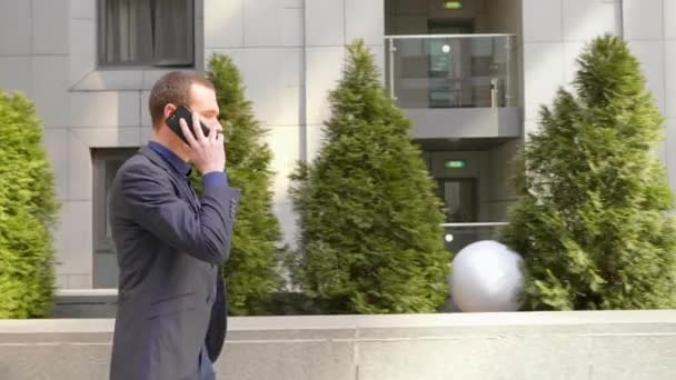 若いビジネスマンが通りを歩き 電話で話す 古典的なスーツを着た男がスマートフォンで会話をリードします 背景に小さな緑色の木 4Kフッテージ — ストック動画