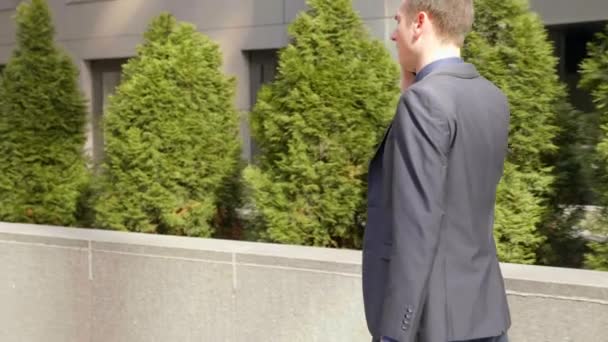 若いビジネスマンが通りを歩き 電話で話す 古典的なスーツを着た男がスマートフォンで会話をリードします 背景に小さな緑色の木 4Kフッテージ — ストック動画