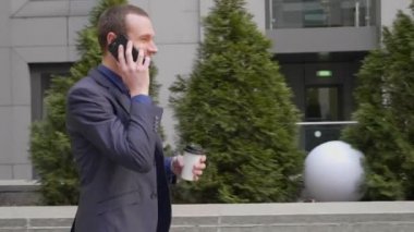 Genç bir işadamı sokakta yürürken mutlu bir telefon görüşmesi iletişim kurar. Elinde bir bardak kahve ile takım elbiseli neşeli bir adam akıllı telefonda konuşurken gülümsüyor. 4k görüntü.