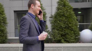 Genç bir işadamı sokakta yürürken mutlu bir telefon görüşmesi iletişim kurar. Elinde bir bardak kahve ile takım elbiseli neşeli bir adam akıllı telefonda konuşurken gülümsüyor. 4k görüntü.