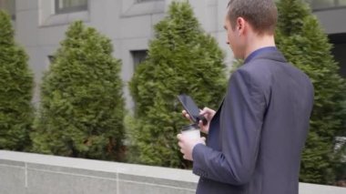 Elinde bir bardak kahveyle caddede yürüyen genç işadamı telefona bir mesaj yazıyor. Klasik takım elbiseli adam kahve içer ve akıllı telefon kullanır. 4k görüntü