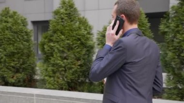 Genç bir işadamı sokakta yürürken mutlu bir telefon görüşmesi iletişim kurar. Elinde bir bardak kahve ile takım elbiseli neşeli bir adam akıllı telefonda konuşurken gülümsüyor. 4k görüntü.