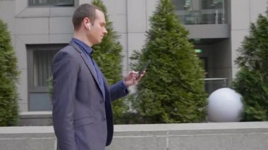 Genç işadamı kablosuz kulaklıkla sokakta yürüyor ve akıllı telefona bir mesaj yazıyor. Klasik takım elbiseli adam kendi telefonunu kullanıyor. Arka planda küçük yeşil ağaçlar var. 4k görüntü.