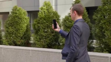 Genç bir işadamı sokakta yürüyor ve görüntülü görüşme hakkında konuşuyor. Klasik takım elbiseli genç bir adam video konferansında bir konuşma açar. Arka planda küçük yeşil ağaçlar. 4k görüntü.