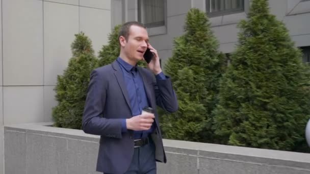 若いビジネスマンが通りを歩き 電話で話す 古典的なスーツを着た男がコーヒーを片手に スマートフォンで会話をリードしている 4Kフッテージ — ストック動画