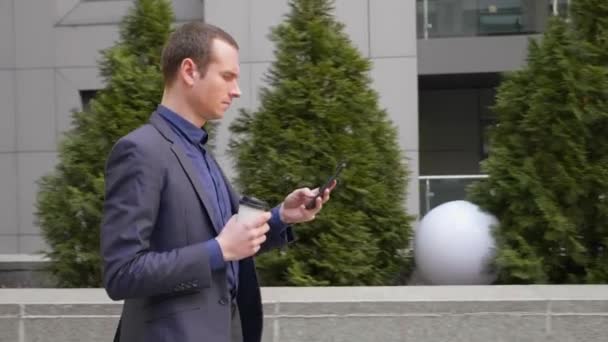 年轻的商人手里拿着一杯咖啡在街上走着 他在电话上写着一条信息 那个穿着经典西装的男人喝咖啡 用的是智能手机 4K镜头 — 图库视频影像