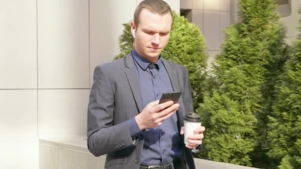 年轻的商人带着无线耳机在街上走来走去 在电话上留言 那个穿着经典西装的男人喝咖啡 用的是智能手机 4K镜头 — 图库视频影像
