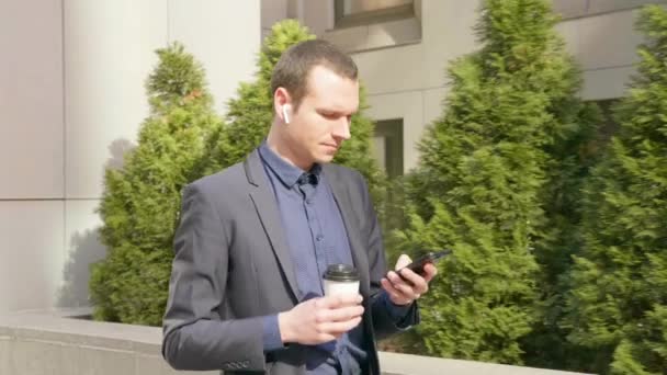 若いビジネスマンが耳の中にワイヤレスイヤフォンで通りを歩いて 電話でメッセージを書きます クラシックなスーツの男はコーヒーを飲み スマートフォンを使用しました 4K映像 — ストック動画