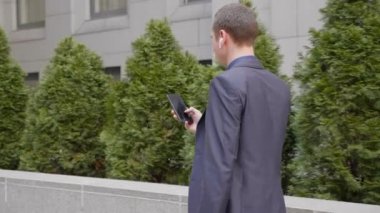 Genç işadamı kablosuz kulaklıkla sokakta yürüyor ve akıllı telefona bir mesaj yazıyor. Klasik takım elbiseli adam kendi telefonunu kullanıyor. Arka planda küçük yeşil ağaçlar var. 4k görüntü.