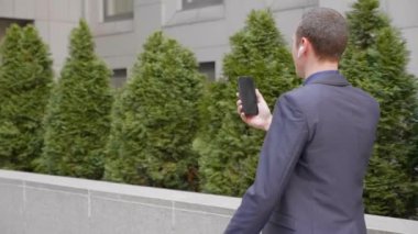 Genç bir işadamı kulaklarında kablosuz kulaklıklarla yürüyor ve akıllı telefondan görüntülü görüşme yapıyor. Video konferansta konuşan takım elbiseli bir adam. 4k görüntü