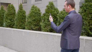 Genç bir işadamı kulaklarında kablosuz kulaklıklarla yürüyor ve akıllı telefondan görüntülü görüşme yapıyor. Video konferansta konuşan takım elbiseli bir adam. 4k görüntü