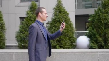 Genç bir işadamı kulaklarında kablosuz kulaklıklarla yürüyor ve akıllı telefondan görüntülü görüşme yapıyor. Video konferansta konuşan takım elbiseli bir adam. 4k görüntü