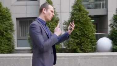 Genç işadamı kablosuz kulaklıkla yürüyor ve akıllı telefondaki bir video görüşmesini agresif bir şekilde yönetiyor. Takım elbiseli bir adam video konferansında hoşnutsuzluğunu gösterir. 4k görüntü