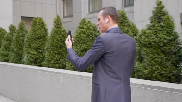 若いビジネスマンがワイヤレスイヤフォンで歩き 積極的にスマートフォンでのビデオ通話についての議論をリードしています 訴訟の男はビデオ会議で彼の不満を示しています 4K映像 — ストック動画