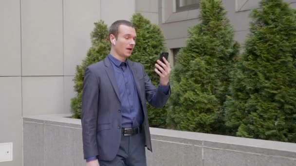 一位年轻的商人带着无线耳机走路 在智能手机上用视频通话 一个穿西装的家伙在视频会议上讲话 — 图库视频影像
