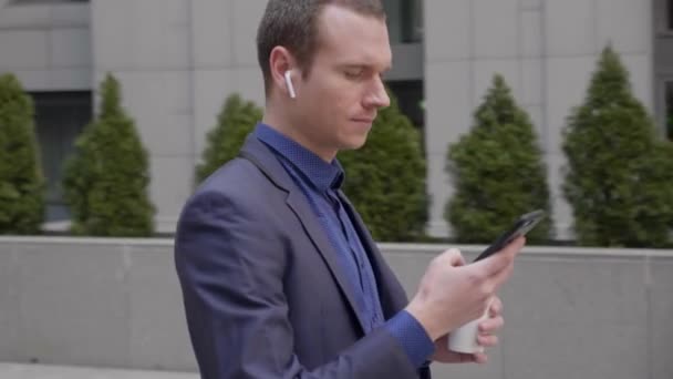 一位耳边装有无线耳机的年轻商人正在智能手机上留言 照相机绕着他转 那个穿经典西装的家伙正拿着一杯咖啡站在大街上 — 图库视频影像