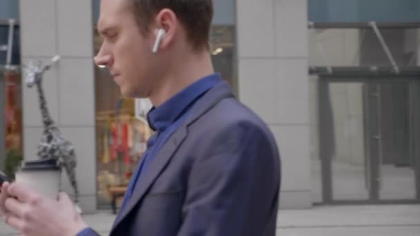 一位耳边装有无线耳机的年轻商人正在智能手机上留言 照相机绕着他转 穿西装的家伙正站在大街上喝咖啡 — 图库视频影像