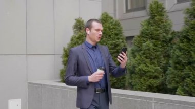 Genç iş adamı kulaklıklarında kablosuz kulaklıkla yürüyor ve akıllı telefondaki video konuşmalarıyla konuşuyor. Takım elbiseli adam, video konferansında konuşuyor ve kahve içiyor. 4k görüntü