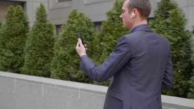 Genç bir işadamı kulaklarında kablosuz kulaklıklarla yürüyor ve akıllı telefondan görüntülü görüşme yapıyor. Video konferansta konuşan takım elbiseli bir adam. 4k görüntü
