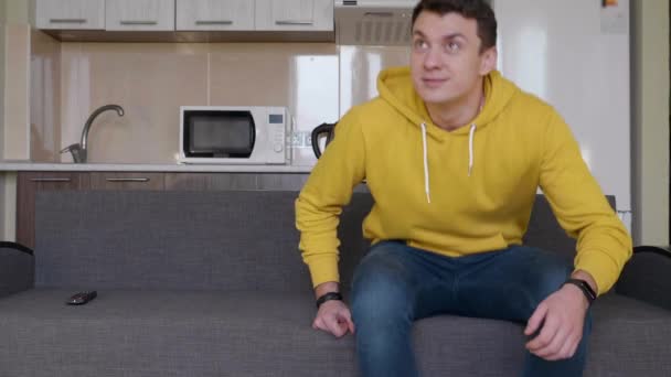 男はソファに座って笑いながら 若い男が立ち上がって立ち去る ソファのテレビのリモコン 背景にキッチン 4Kフッテージ — ストック動画