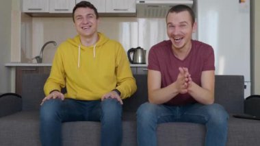 İki adam kanepede oturuyor ve gülümsüyor. Genç adamlar sert güler, arkadaşlarından biri alkışlar ve ellerini sallar. Arka planda mutfak var. 4k görüntü.