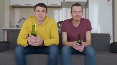 İki adam kanepede oturup televizyonda bir futbol maçı izliyor ve ellerinde bira şişeleri tutuyorlar. İki genç adam sakince kanepede bir spor maçı izliyor. Backgroung'da bir mutfak. 4k görüntü.
