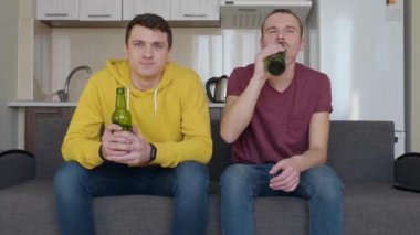 İki adam kanepede oturup televizyonda bir futbol maçı izleyip bira içiyor. İki genç adam sakince kanepede bir spor maçı izliyor. Backgroung'da bir mutfak. 4k görüntü.