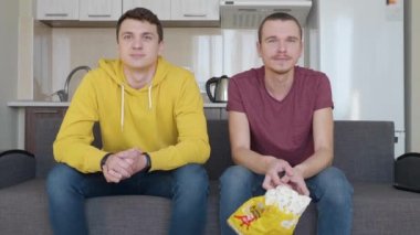 İki adam bir futbol maçı izliyor. Genç çocuklar kanepede oturuyor ve tv izliyor, bir arkadaş aperatifler bir paket yakalar. Arka plandaki mutfak. 4k görüntü.