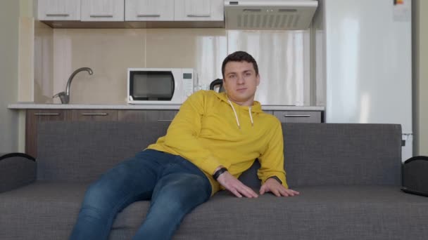 男がソファで休んでいる 座ってリクライニングし ソファをノックする若い男 背景にキッチン 4Kフッテージ — ストック動画