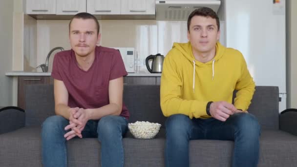 坐在沙发上的两个人用爆米花碗在电视上观看一场足球比赛 两个年轻人平静地看一场体育比赛 背景上的厨房 — 图库视频影像