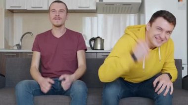 Kanepede oturan iki adam tv'de bir futbol maçı izlemek ve en sevdikleri takımın zaferinin tadını çıkarın. Genç çocuklar spor oyunu lehine ve sevinç için bağırmak vardır. Arka plandaki mutfak. 4k görüntü.